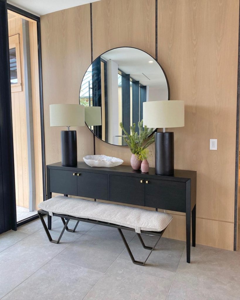 Modern vibe for the foyer or entryway. Includes wooden wall panels, large round mirrors, and black console table with lamps and other accessories. 